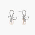 Silver bow-shaped stud earrings with baguette crystals and dangling white freshwater pearls on cream background