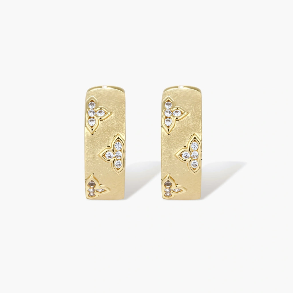 Gold-plated hoop earrings with textured surface and raised floral motifs set with clear zirconia stones on cream background.