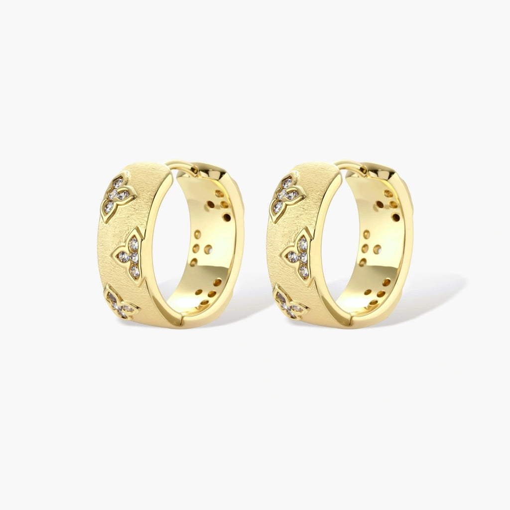 Gold-plated hoop earrings with textured surface and raised floral motifs set with clear zirconia stones on cream background.