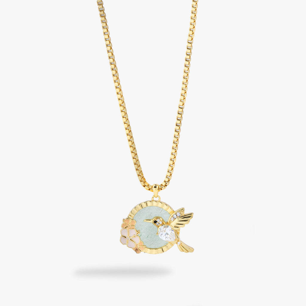 A gold chain necklace with a circular pendant featuring a light green enamel center, a gold hummingbird charm with a clear crystal, and small pale pink floral accents, shown on a light gray background.
