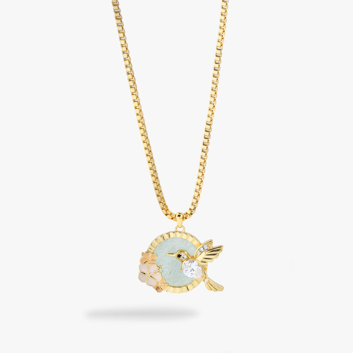 A gold chain necklace with a circular pendant featuring a light green enamel center, a gold hummingbird charm with a clear crystal, and small pale pink floral accents, shown on a light gray background.